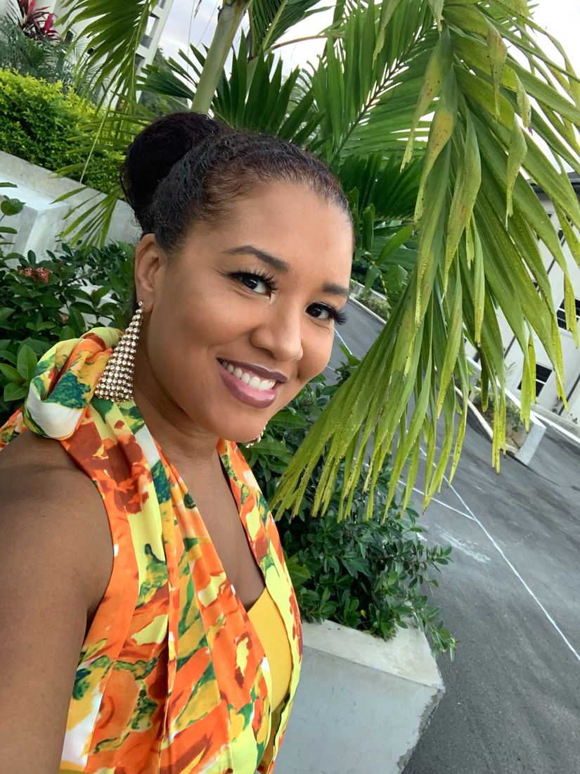 Dr. Tanika Lawrence smiling under palm fronds in Jamaica, wearing an orange and yellow print