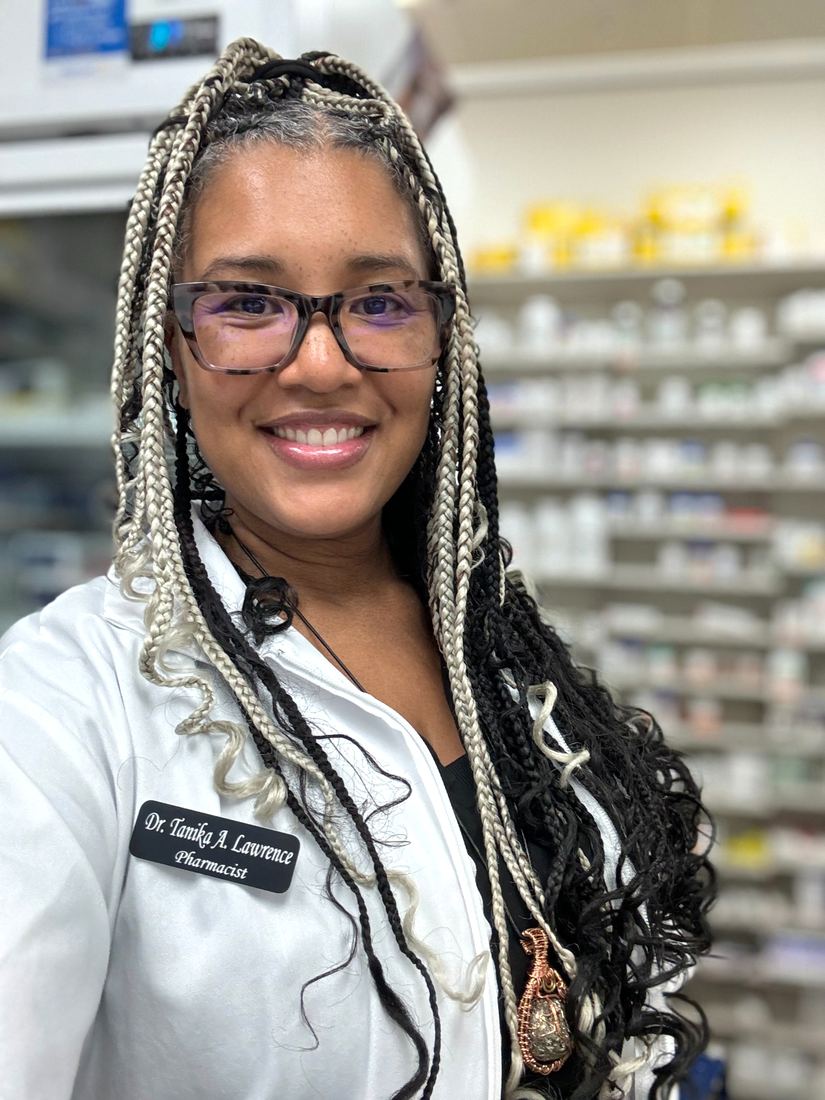 Dr. Tanika Lawrence smiling in her pharmacist’s white coat, with long braids and glasses